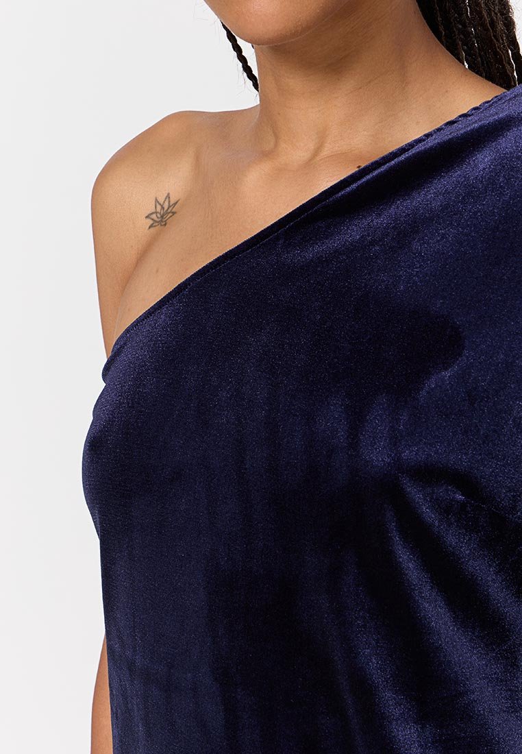 Frau trägt ein dunkelblaues Samt-Top mit einem Schulterträger, das ein kleines Tattoo eines Blattes auf ihrer linken Schulter zeigt.