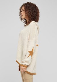 Knit hoodie in cream with a relaxed fit, featuring a large brown star pattern on one side and ribbed cuffs and hem in matching brown.