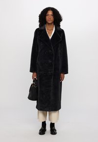 Long, dark fur coat with a double-breasted front, large collar, and black buttons, paired with a black handbag and cream-colored trousers.