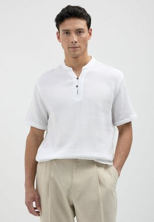 Chemise blanche à manches courtes en tissu texturé, décolleté en V et patte de boutonnage à deux boutons. Associée à un pantalon beige.