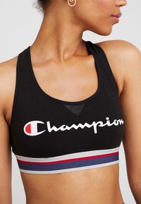 Sujeción deportiva negra con una banda ancha de rayas grises, azul marino y rojas en la parte inferior, con un logo blanco de "Champion" y un panel de malla.