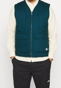 Tealfärgad väst av vävt tyg, med en dragkedja framtill, två stora fickor med lappar och en varumärkesetikett på fållen.