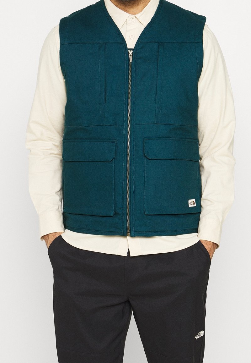 Tealfärgad väst av vävt tyg, med en dragkedja framtill, två stora fickor med lappar och en varumärkesetikett på fållen.