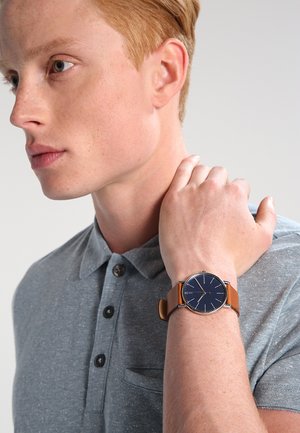 Jeune homme en polo gris avec cheveux courts roux portant une montre bracelet en cuir marron avec un cadran bleu foncé au poignet gauche.