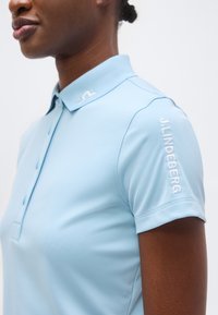 Γυναίκα που φοράει ανοιχτό γαλάζιο πόλο με γιακά και τη λέξη "J.LINDEBERG" κεντημένη στο δεξί μανίκι.