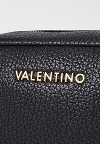 Juodas tekstūruotas odos krepšys su auksinės spalvos "VALENTINO" logotipu priekyje. Lygus uždarymas su šiek tiek pakeltais raštais.