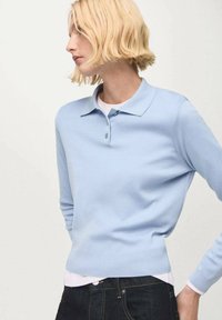Hellblauer, enganliegender, langärmliger Poloshirt-Pullover mit Kragen, der eine glatte Textur aufweist und einen einzelnen Knopf am Hals hat, über einem weißen Hemd getragen.
