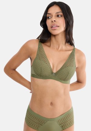 Frau trägt olivgrünen Spitzen-BH und passende Unterwäsche, posiert mit den Händen hinter dem Rücken vor einem einfarbigen, hellen Hintergrund.