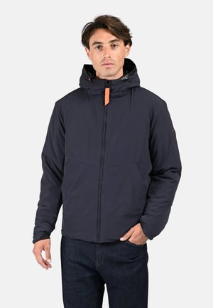 Homme portant une veste à capuche bleu marine avec un zip orange, se tenant avec une main dans la poche et une expression neutre sur un fond clair.