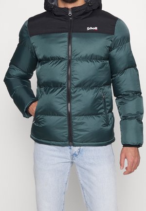 Mężczyzna w zielonej kurtce puchowej z czarnymi panelami na ramionach, zapinanej na zamek, z kieszeniami po bokach, w jasnoniebieskich dżinsach.