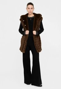 Brązowa kamizelka z sztucznego futra z szerokim kołnierzem, zapinana na guziki, z teksturowanym wzorem, noszona na czarnej bluzce z długim rękawem i spodniach.