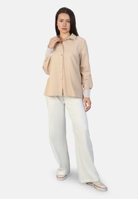 Beige long-sleeve shirt with button closure and textured cuffs, paired with white wide-leg pants and casual sneakers. Plain background.