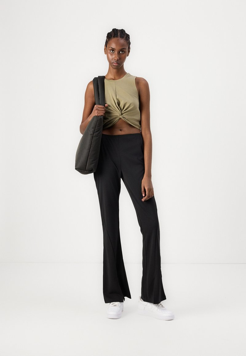 Olivgrünes Crop-Top mit gedrehtem Vorderteil, schwarze Schlaghosen und schwarze Umhängetasche. Weiße Sneaker vervollständigen den Look.