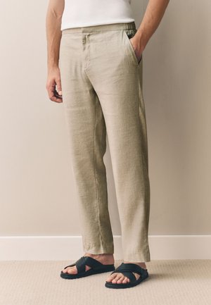 Mężczyzna ubrany w beżowe lniane spodnie, białą koszulę i czarne, krzyżowane klapki, stojący z jedną ręką w kieszeni na tle neutralnej ściany.