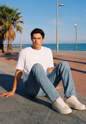 Młody mężczyzna w białej koszulce i dżinsach siedzi na nadmorskiej promenadzie w pobliżu palm pod czystym, niebieskim niebem.