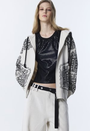 Jonge vrouw die een witte hoodie draagt met zwarte abstracte patronen, een zwart topje met pailletten, witte broek en een zwarte riem met een hangende band.