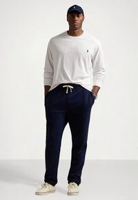 Hosszú ujjú fehér póló, sötétkék melegítőnadrág húzózsinórral, és bézs sportcipők; további sötétkék logós sapka. Laza öltözék, egyszerű dizájn.