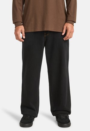 Zwarte denim jeans met een relaxte pasvorm, voorzien van oranje stiksels en zakken, gecombineerd met zwarte schoenen en een bruine blouse met lange mouwen.