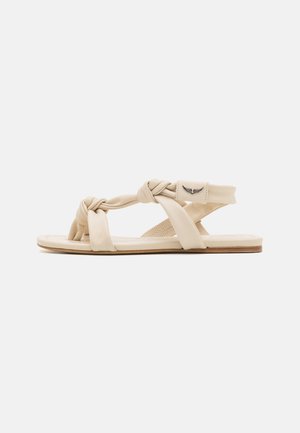 Beige flad sandal med snoede stropper og åben tå, med en lille vingeemblem på ankelstroppen mod en hvid baggrund.