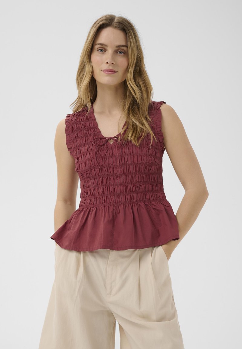 Femme aux longs cheveux blonds portant un top smocké sans manches couleur rouille et un pantalon beige taille haute, mains dans les poches, fond neutre.