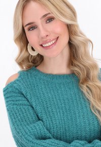 Teal színű, ribbelt textúrájú kötött pulóver, nyitott vállú kialakítással. A modellen kerek, fonott fülbevalók láthatók. Puha és kényelmes megjelenés.
