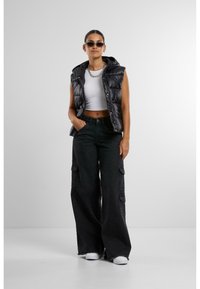 Gilet rembourré noir avec capuche sur un crop top blanc, associé à un pantalon cargo noir à jambes larges avec poches latérales et des baskets blanches.