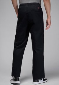 Black elastic waistband pants with a straight leg design, featuring a rear pocket and a small logo patch. Worn with black and white sneakers.