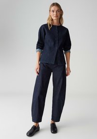 Dunkles Denim-Outfit mit einem Oberteil zum Knöpfen und hochgekrempelten Ärmeln sowie weitem Hosenbein. Der Stoff hat eine glatte Textur und klare Linien.