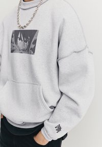 Grå hættetrøje med en frontlomme, ribbet krave og lange ærmer. Har et grafisk tryk og et "DRA" mærke på ærmekanten.