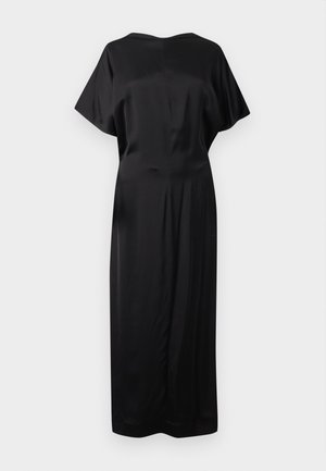 Žena stoji s rukom na kuku, nosi crnu midi haljinu s kratkim rukavima i bijele cipele šiljastih vrhova na bijeloj pozadini.