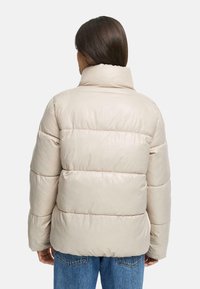 Beige gewatteerde jas met een hoge kraag, voorzien van horizontale gestikte panelen en een glanzende textuur, gecombineerd met denim jeans.