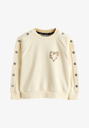Ung kvinde med langt brunt hår bærer en cremefarvet sweatshirt med et blomsterhjerte og teksten "All Saints" på ryggen, kombineret med falmede sorte jeans.
