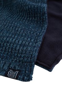 Stickad scarf i mörkblå och tealtoner med texturerad mönster; mjukt fleecetyg på baksidan och en etikett med en stiliserad "BR"-logotyp.