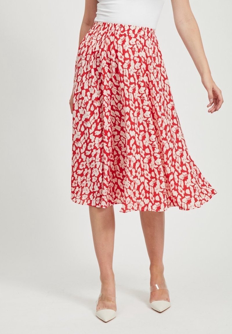 Femme portant une jupe midi rouge à motifs floraux blancs, associée à des talons blancs à bouts pointus et un haut blanc sans manches.