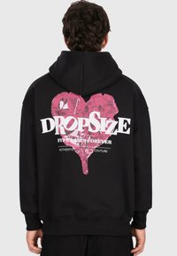 Person with dark curly hair wearing black hoodie with large pink heart and white "DROPSIZE" text on the back.