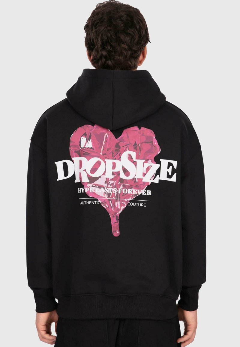 Personne avec des cheveux bouclés foncés portant un sweat à capuche noir avec un grand cœur rose et le texte blanc "DROPSIZE" dans le dos.