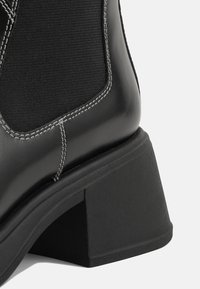 Must black leather ankle boot with elastic fabric panel, square block heel, contrasting white stitching, and smooth texture.