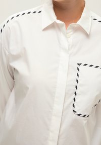 Weißes Hemd mit spitzem Kragen, Brusttasche mit schwarz-weißem diagonalen Streifenmuster und kontrastierenden Besätzen an den Schultern.