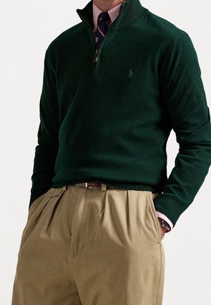 Homme portant un pull à fermeture éclair vert foncé sur une chemise rose et une cravate bleu marine, associé à un pantalon beige plissé, les mains dans les poches.