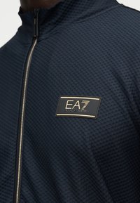 Ναυτικό μπουφάν με φερμουάρ, κατασκευασμένο από υφασμά με υφή, που διαθέτει σημάδι λογότυπου με χρυσή λεπτομέρεια στο στήθος και λεπτό σχέδιο για το γιακά.