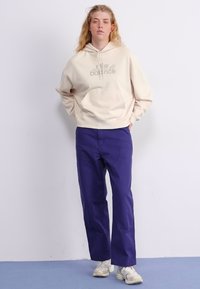 Beige hettegenser med en lomme foran og logo, sammen med løstsittende lilla bukser og hvite sneakers på en ensfarget bakgrunn.