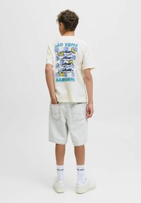 Beige katoenen t-shirt met een grafisch ontwerp van vissen met citroenen, "SÃO TOMÉ" en de tekst "SARDINHA" op de achterkant. Gepaard met lichte denim shorts.