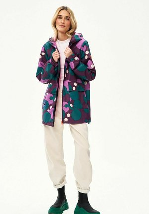 Kobieta ubrana w kolorowy płaszcz przeciwdeszczowy w geometryczne wzory w kolorach morskim, fioletowym i różowym, w połączeniu z białymi spodniami i czarnymi butami z zielonymi podeszwami.