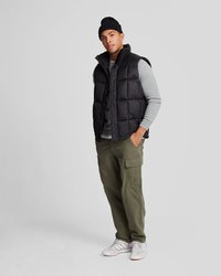 Czarna pikowana kamizelka na szarym swetrze, oliwkowe spodnie cargo i białe sneakersy. Model stoi na białym tle, nosząc czarną czapkę beanie.