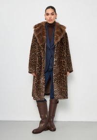 Cappotto con stampa leopardata e colletto in finta pelliccia marrone, indossato sopra un dolcevita scuro e un completo blu, abbinato a stivali marroni fino al ginocchio.