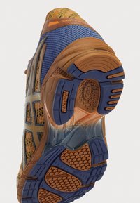 Sneaker med en texturerad brun och blå ovandel, som har nät- och gummimaterial. Yttersulan har ett bikake-mönster och solida greppande element.