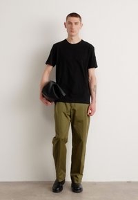 Black cotton t-shirt, olive green trousers with a straight fit, and black leather bag held in one hand. Minimalist design, neutral colors.