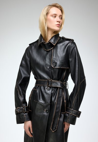 Mustast nahast trench coat, millel on vööga talje, topelt-ketas, suur krae ja kontrastsete õmbluse detailidega.