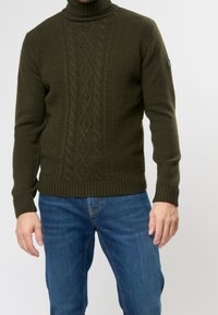 Pull à col roulé vert foncé côtelé avec un motif torsadé texturé et des poignets ajustés, associé à un jean bleu.