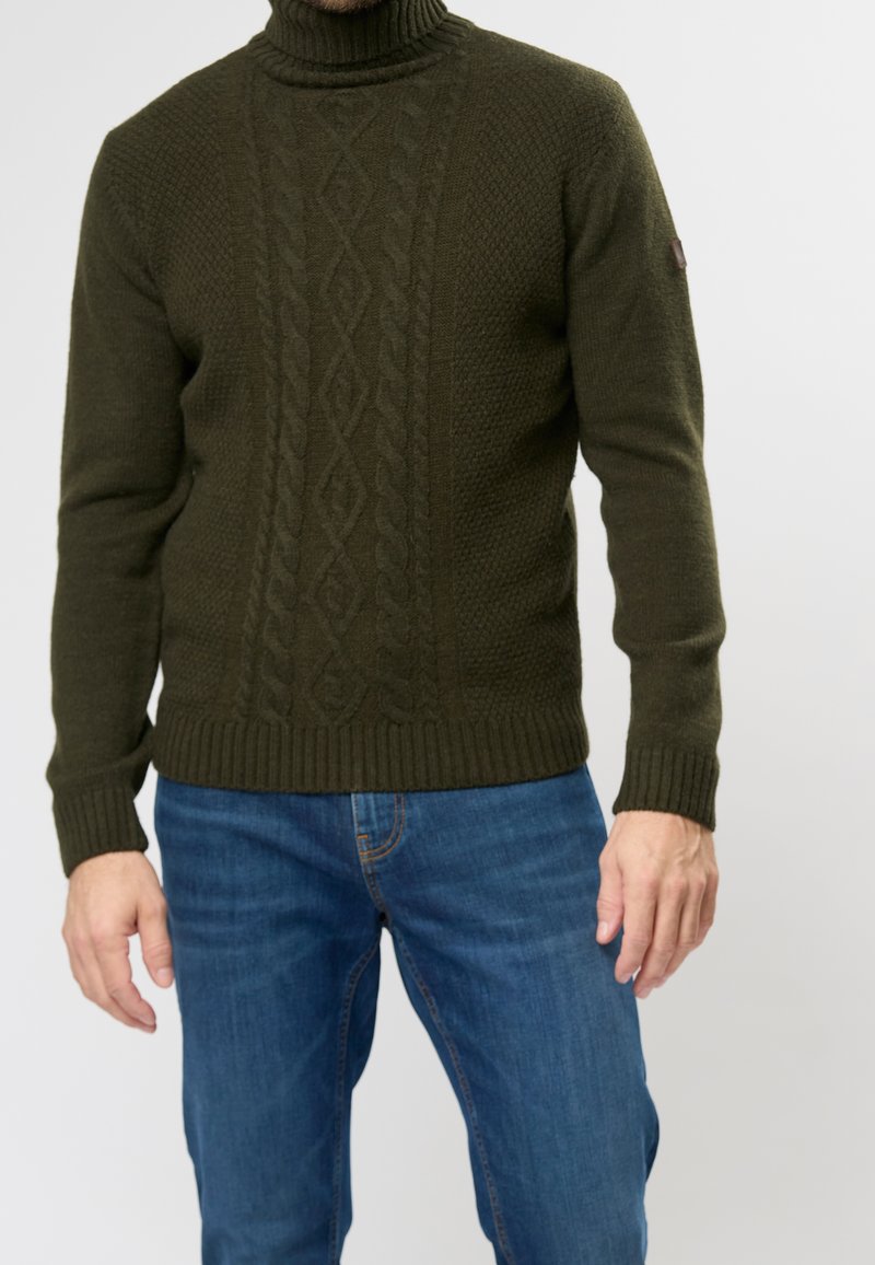 Pull à col roulé vert foncé côtelé avec un motif torsadé texturé et des poignets ajustés, associé à un jean bleu.